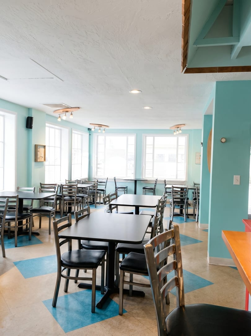 Bright, empty restaurant dining room with blue walls, large windows, and black tables with metal chairs.
